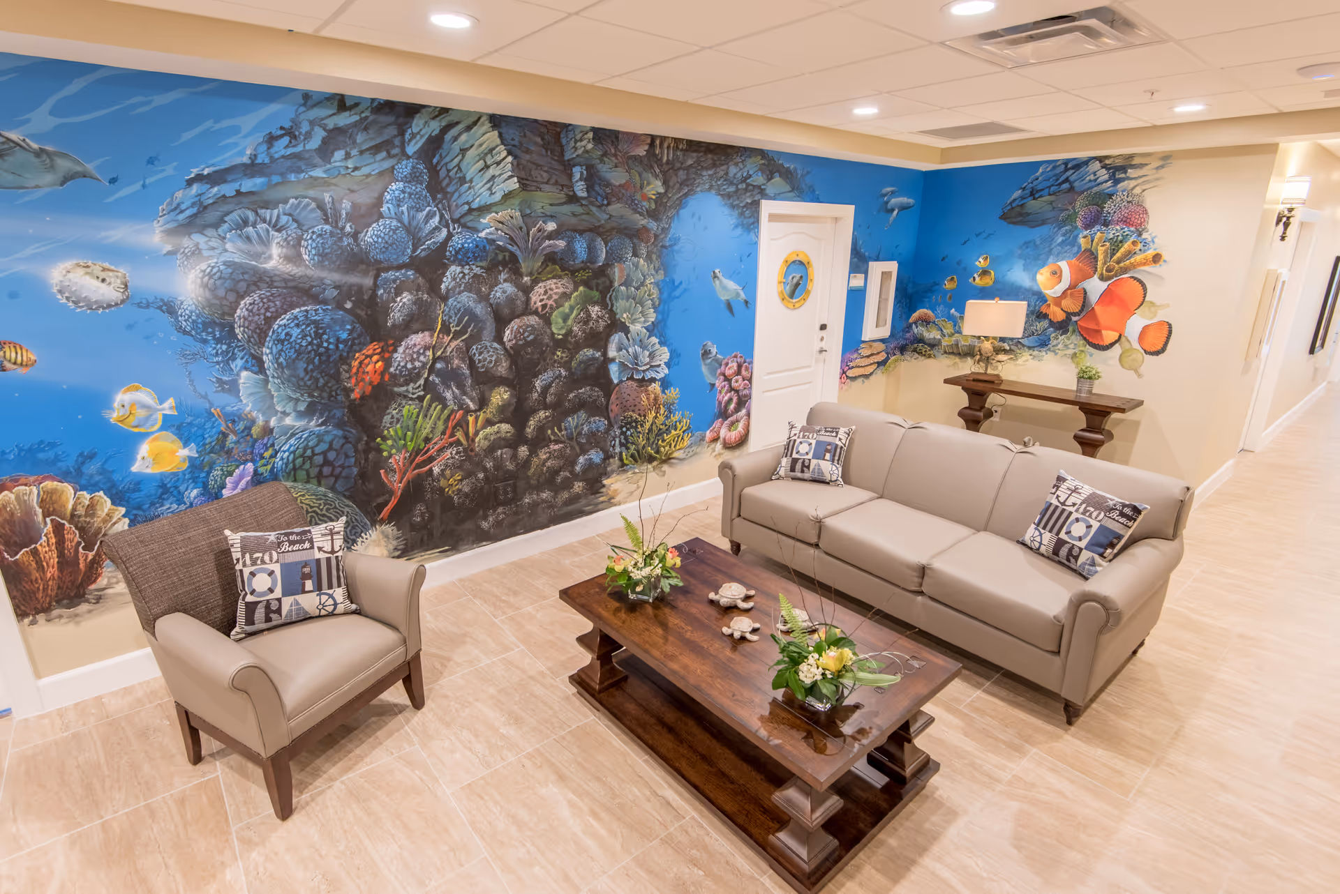 A cozy living room area with a beige leather sofa and armchair, both adorned with decorative pillows. A wooden coffee table with floral arrangements sits in the center. The walls feature a vibrant underwater mural depicting coral reefs, fish, and sea creatures, creating a lively and colorful atmosphere.