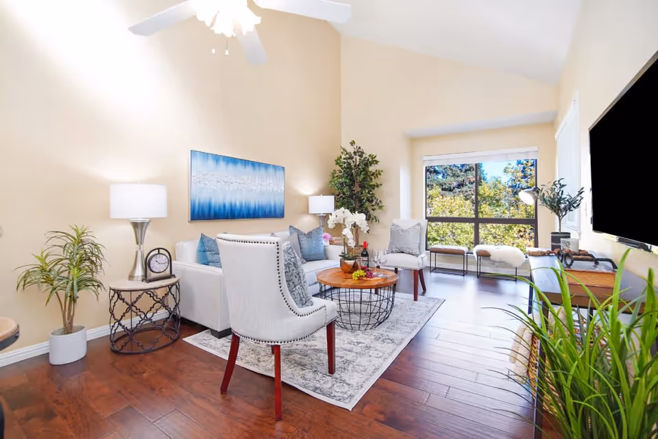 Bright and airy living room with high vaulted ceilings, a ceiling fan, and large window showing greenery outside. The room features a white sofa with blue cushions, two upholstered chairs, a round wooden coffee table with a wireframe base, a side table with a lamp and clock, several potted plants, a wall-mounted flat-screen TV, and decorative artwork on the wall.