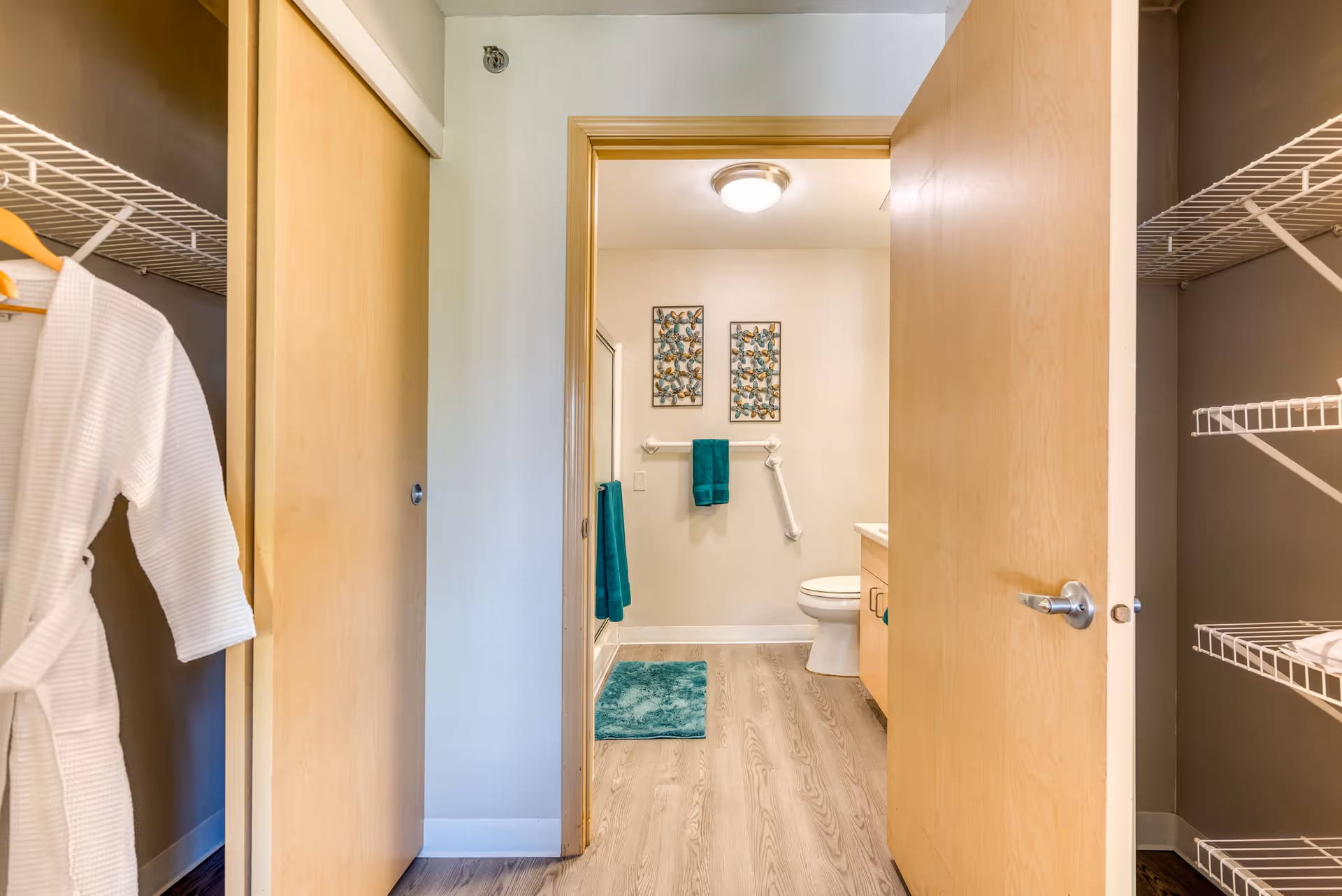 View through an open doorway into a bathroom with light wood flooring, a toilet, a vanity with a sink, teal towels hanging on the wall, and two decorative framed artworks. On either side of the doorway are closets with wire shelving; the left closet has a white robe hanging.