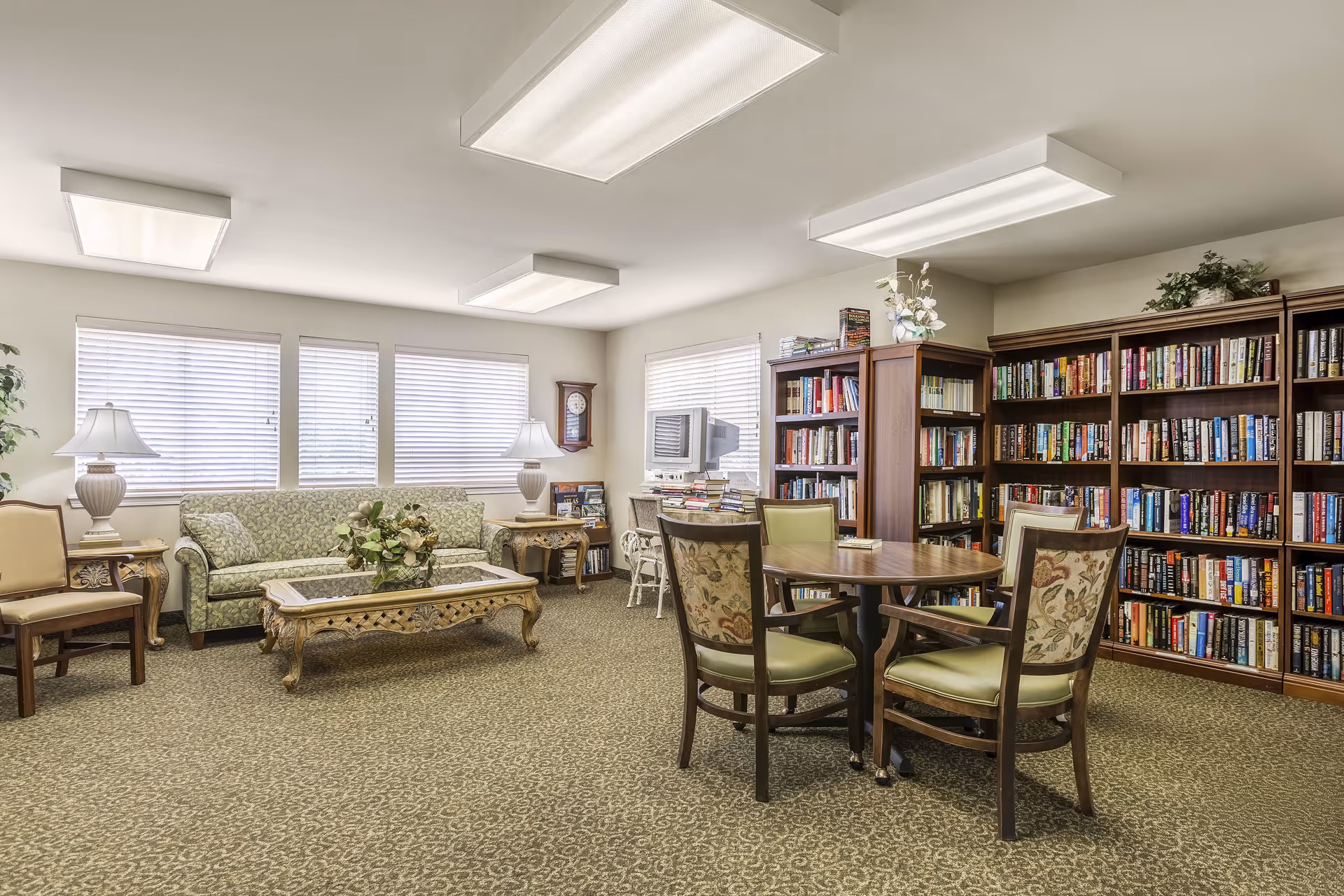 A cozy senior living community library and lounge area with a patterned carpet, a green floral sofa, two side tables with lamps, a wooden coffee table with a floral centerpiece, a round wooden table with four chairs, and multiple bookshelves filled with books. There is a wall clock and a small TV on a stand near the windows with blinds.