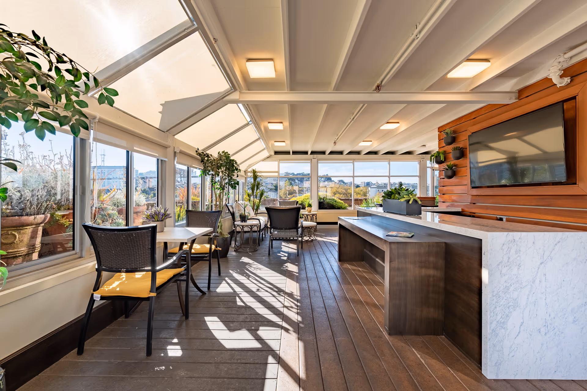 Bright indoor lounge area with large windows and skylights allowing natural light to fill the space. The room features several chairs with yellow cushions, small tables, potted plants, a long marble countertop with a wooden base, and a wall-mounted flat screen TV on a wooden accent wall.