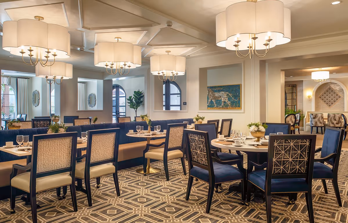 Elegant dining room with multiple tables and chairs arranged neatly. The room features large, decorative chandeliers hanging from a coffered ceiling, patterned carpet flooring, and artwork on the walls. Tables are set with white dishes, glassware, and small floral centerpieces.