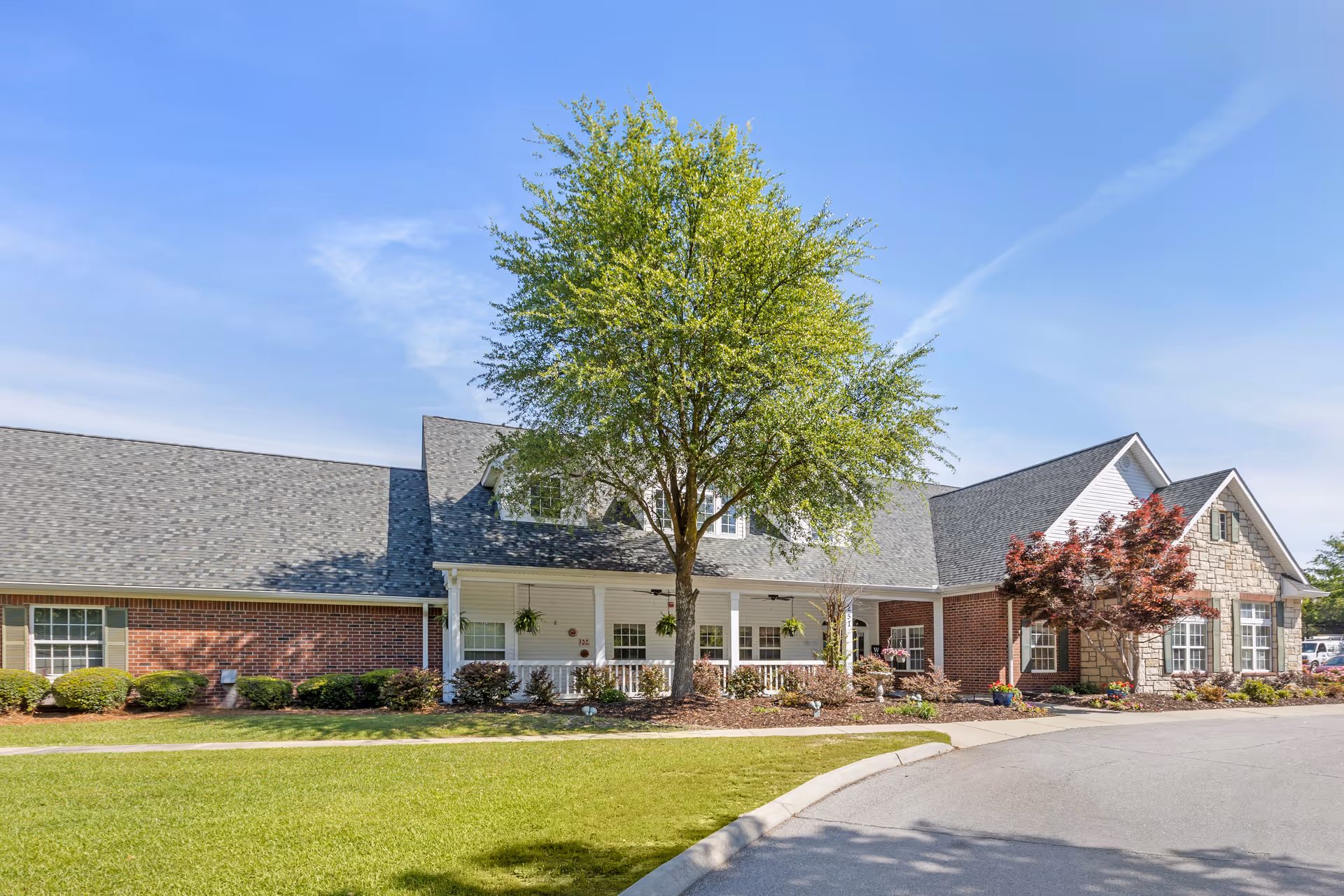 Exterior view of a single-story senior living facility building with a mix of brick and stone facade, a large tree in front, well-maintained landscaping, and a clear blue sky.