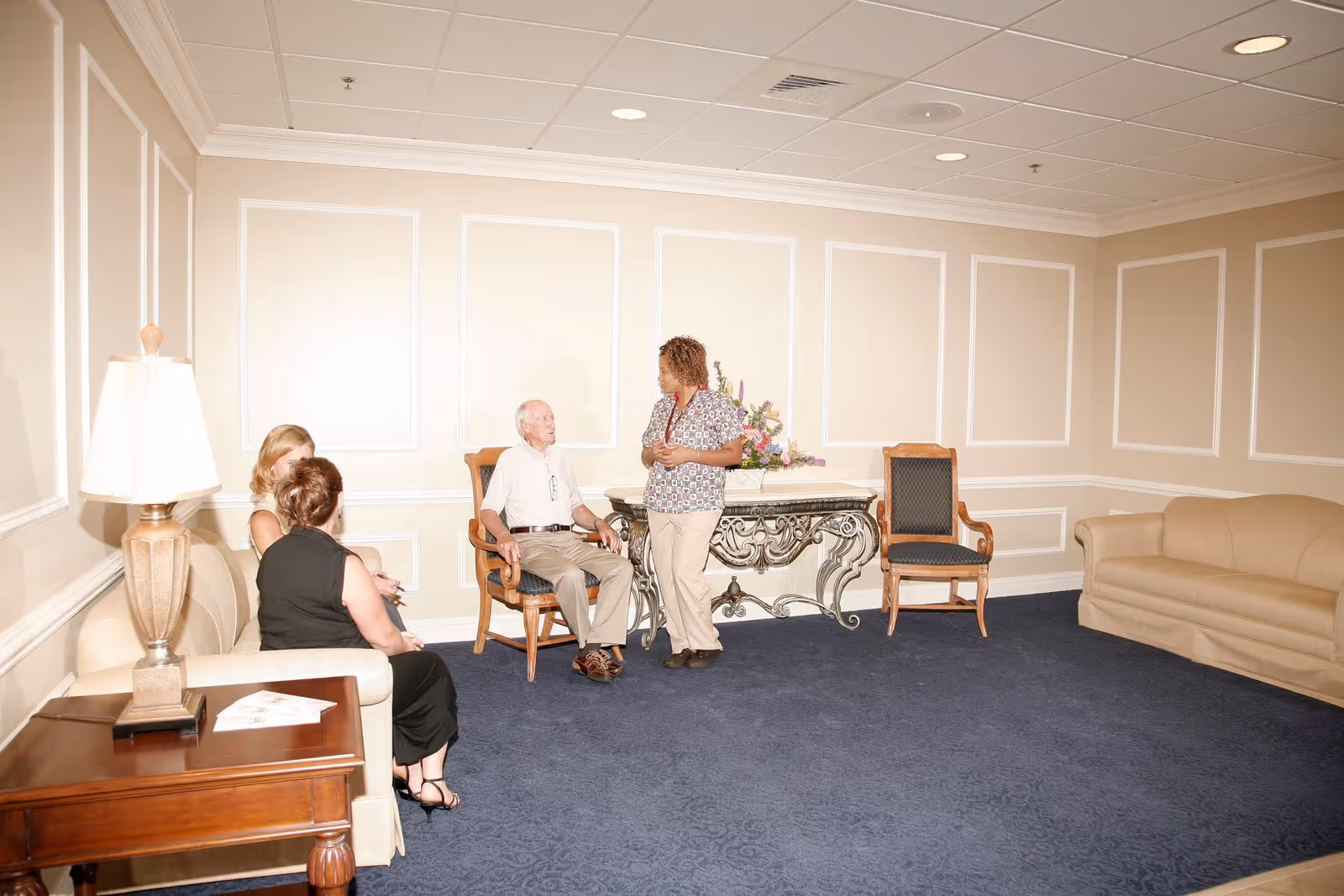 A group of four people in a well-lit room with beige walls and blue carpet. Two women are seated on a beige couch on the left, an elderly man is seated on a wooden chair in the center, and a woman in a patterned top and beige pants is standing and talking to the elderly man. There is a decorative table with a floral arrangement behind them and another wooden chair next to it. A wooden side table with a lamp and some papers is visible in the foreground.