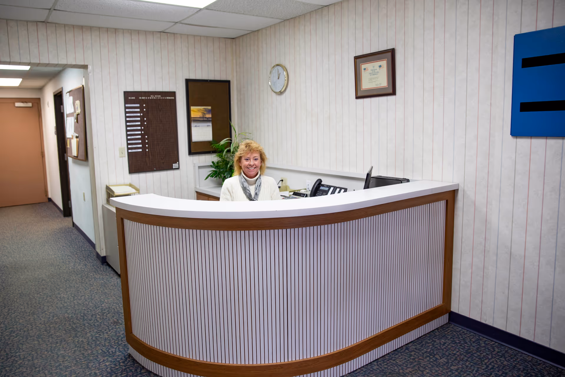 A woman sitting behind a curved reception desk in an office area with beige striped wallpaper, a clock, framed certificate, and bulletin boards on the wall.