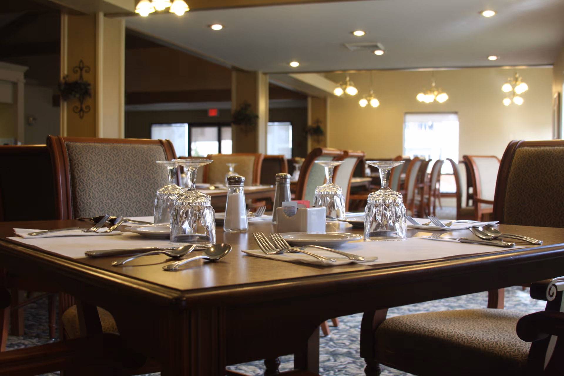 A dining room in a retirement facility with tables set for a meal, including upside-down wine glasses, silverware, salt and pepper shakers, and napkins. The room has upholstered chairs and soft lighting from ceiling fixtures.