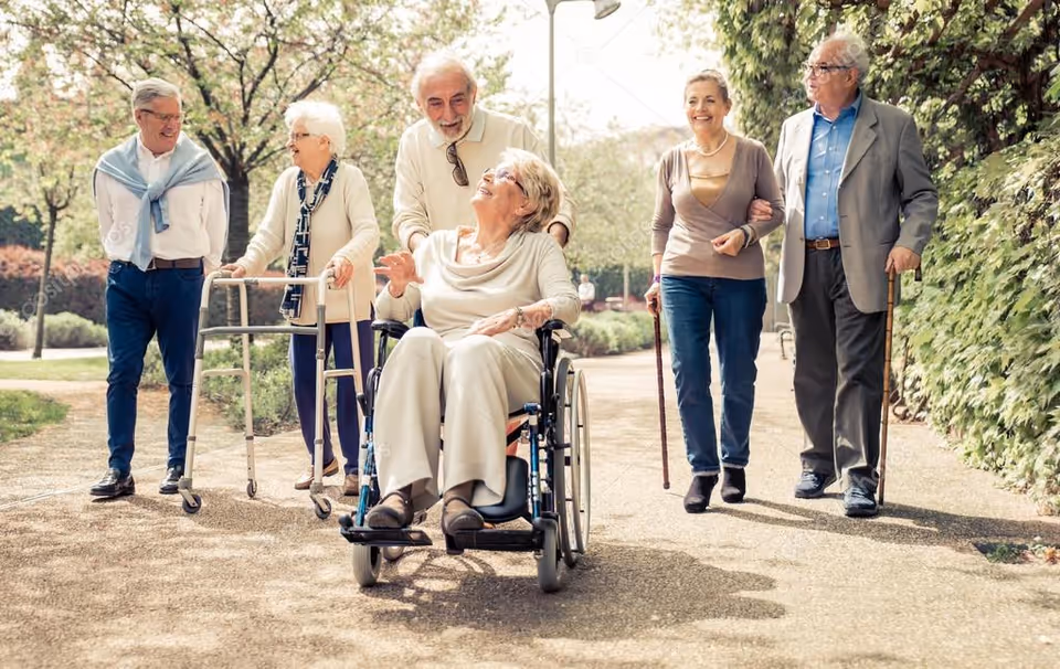 A group of six elderly people enjoying a walk outdoors on a sunny day. One woman is in a wheelchair being pushed by a man, another woman uses a walker, and another man uses a cane. They are all smiling and appear to be engaged in conversation while walking on a paved path surrounded by greenery and trees.