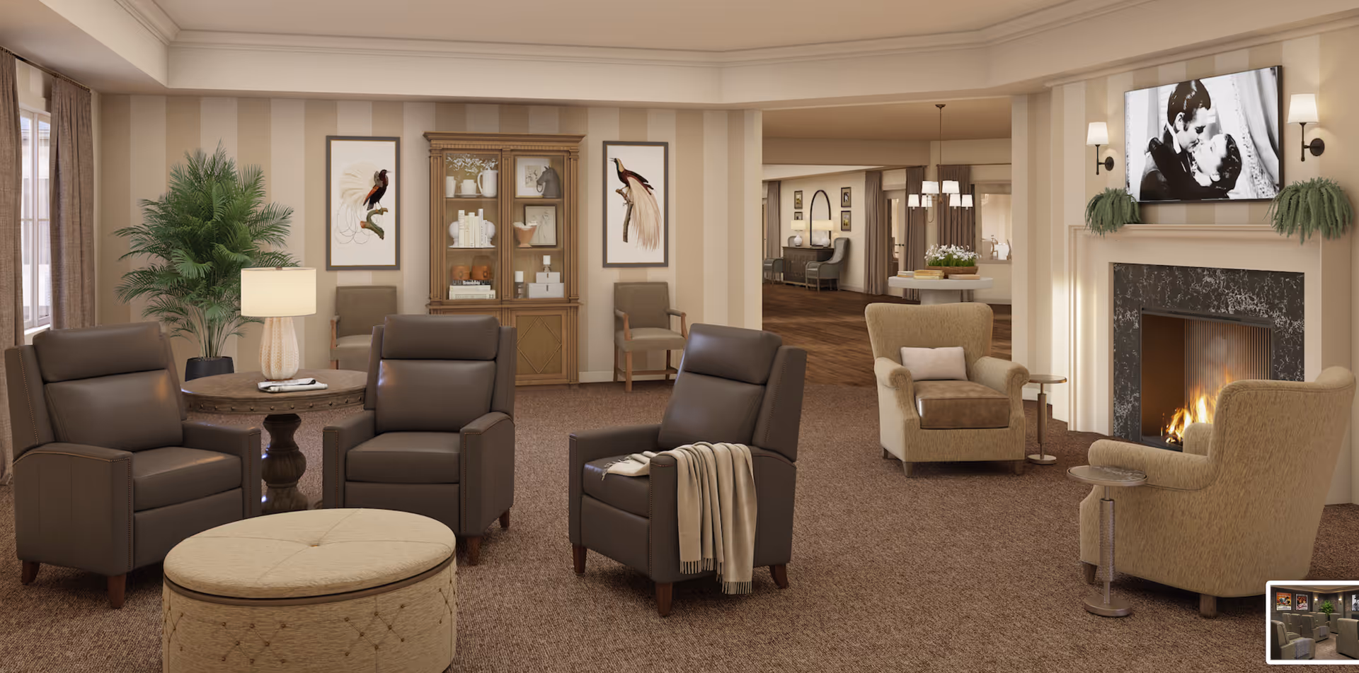 A cozy senior living facility common area with comfortable leather and fabric armchairs arranged around a round ottoman. There is a round wooden side table with a lamp and a large potted plant next to it. The room features a fireplace with a black and white movie playing on a mounted TV above it, flanked by two wall sconces and green plants. The walls have beige vertical stripes and framed bird artwork. In the background, there is a wooden cabinet with decorative items and an open view into another room with more seating and lighting fixtures.