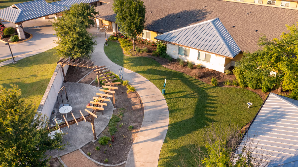 Aerial view of an outdoor garden area at The Waterford at Round Rock featuring a curved concrete pathway, a pergola structure with a small round table and chairs underneath, surrounded by green grass, trees, and landscaped plants near building structures with metal roofs.