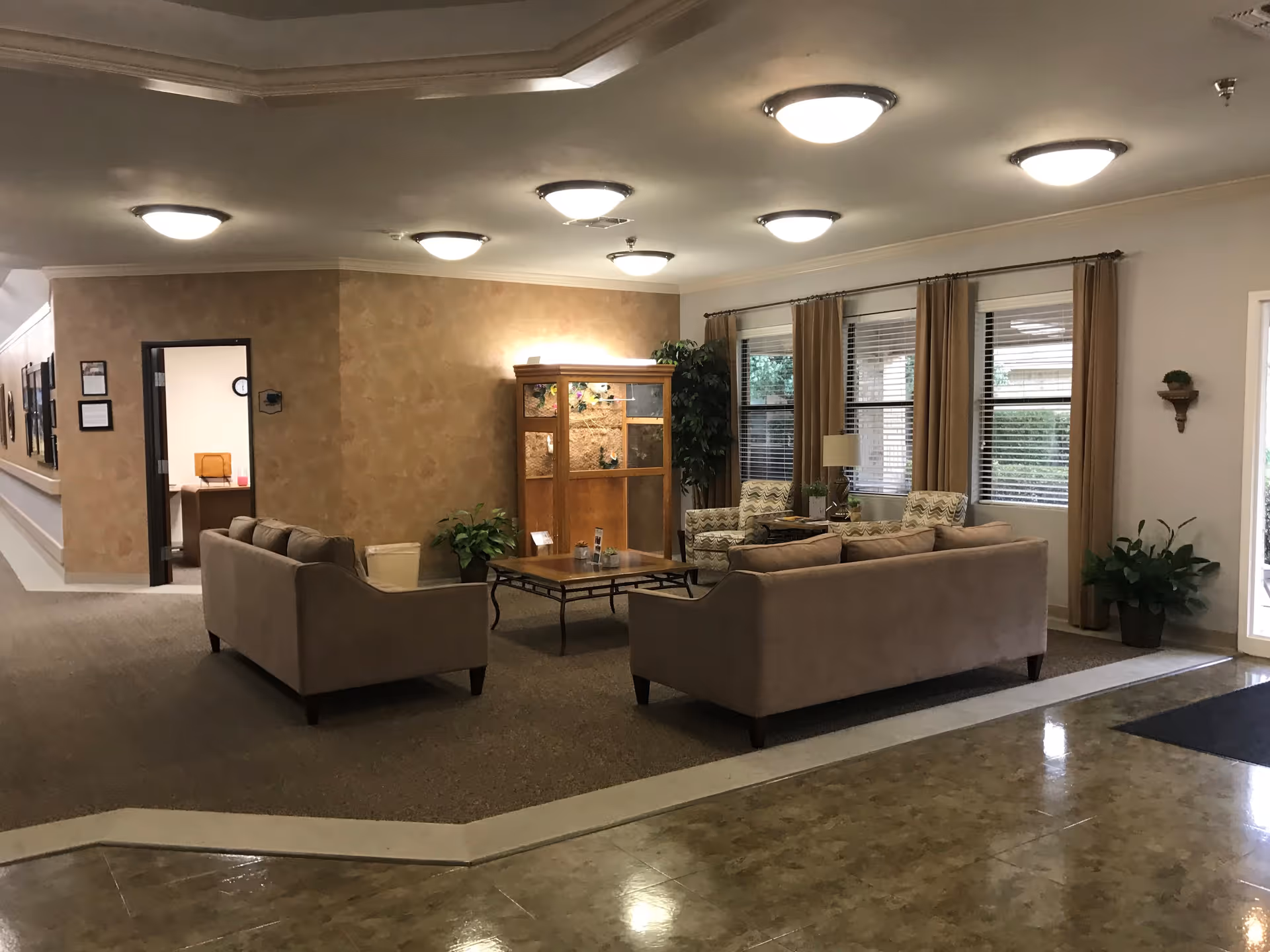 Comfortable lounge area with sofas, armchairs, a coffee table and large windows in a nursing home common room.