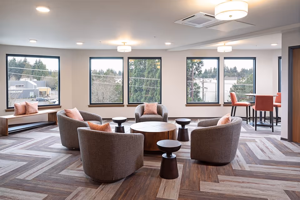 A bright and modern lounge area with four gray armchairs arranged around a round wooden coffee table and three small black side tables. The room has large windows showing trees and buildings outside, a bench with pink cushions under one window, and a small dining table with four coral-colored chairs near the corner. The floor has a patterned wood design, and the ceiling has recessed lighting and two hanging light fixtures.