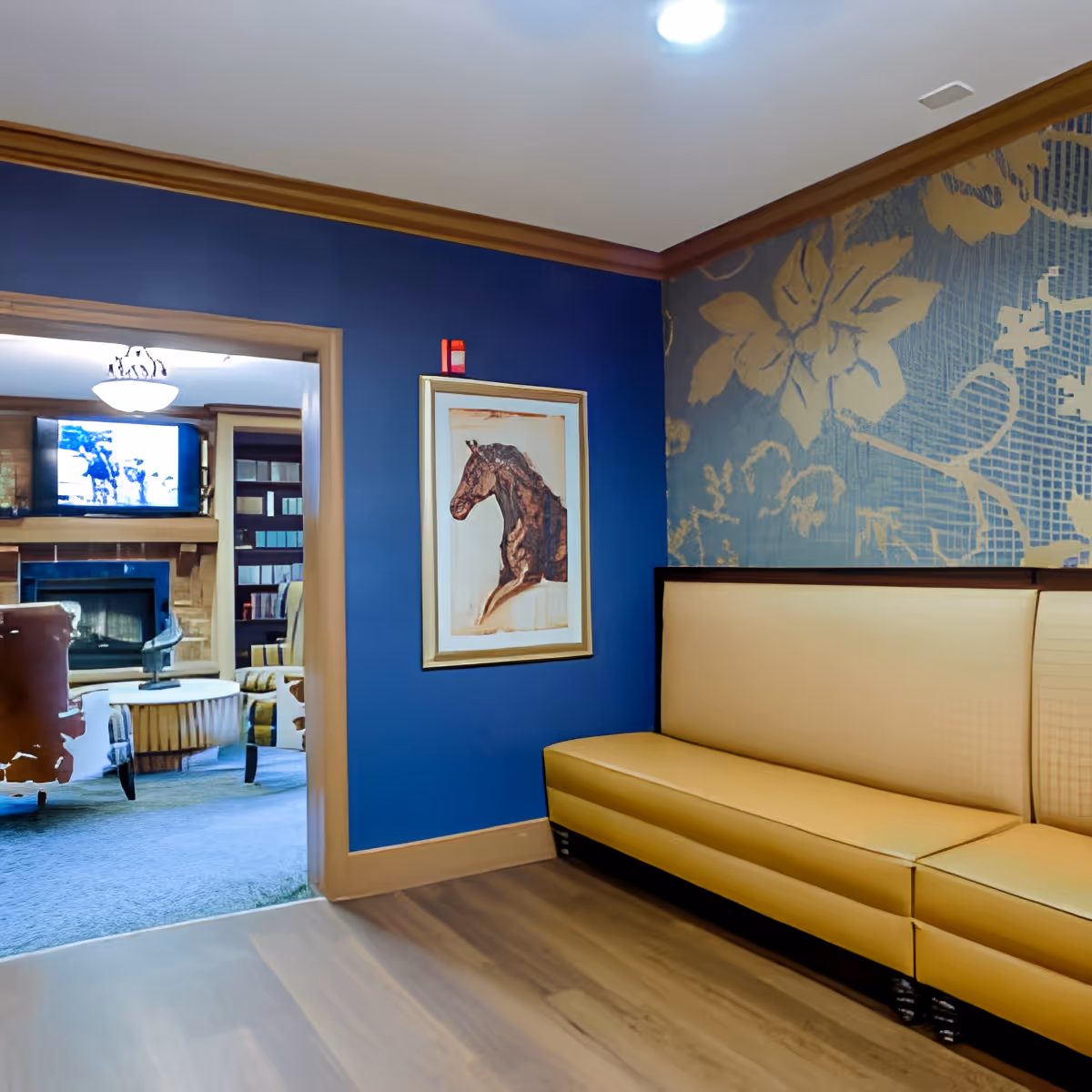 Interior view of a senior living facility lounge area with a blue wall featuring a framed horse artwork, a beige cushioned bench, and a doorway leading to another room with armchairs, a fireplace, and a television.