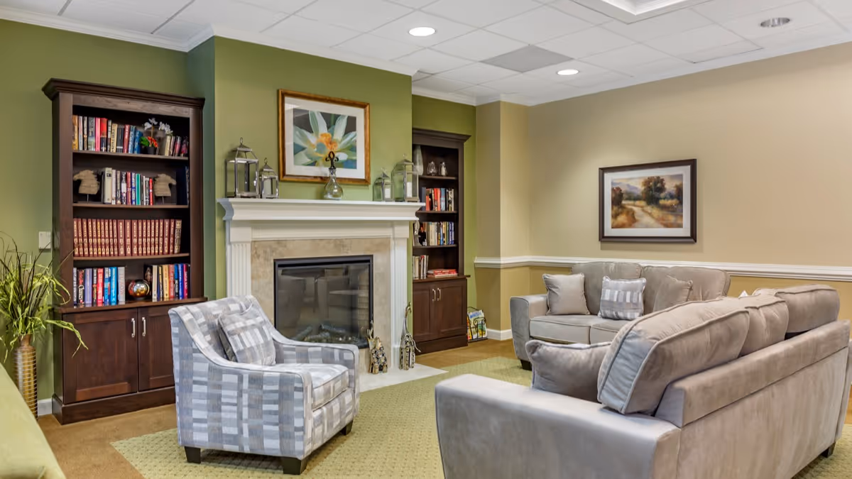 A cozy living room with beige walls and green accent wall featuring a fireplace in the center. On either side of the fireplace are dark wooden bookshelves filled with books and decorative items. In front of the fireplace is a patterned armchair, and to the right are two beige sofas with cushions. A framed landscape painting hangs on the wall above one sofa, and the room is carpeted with a light green rug.