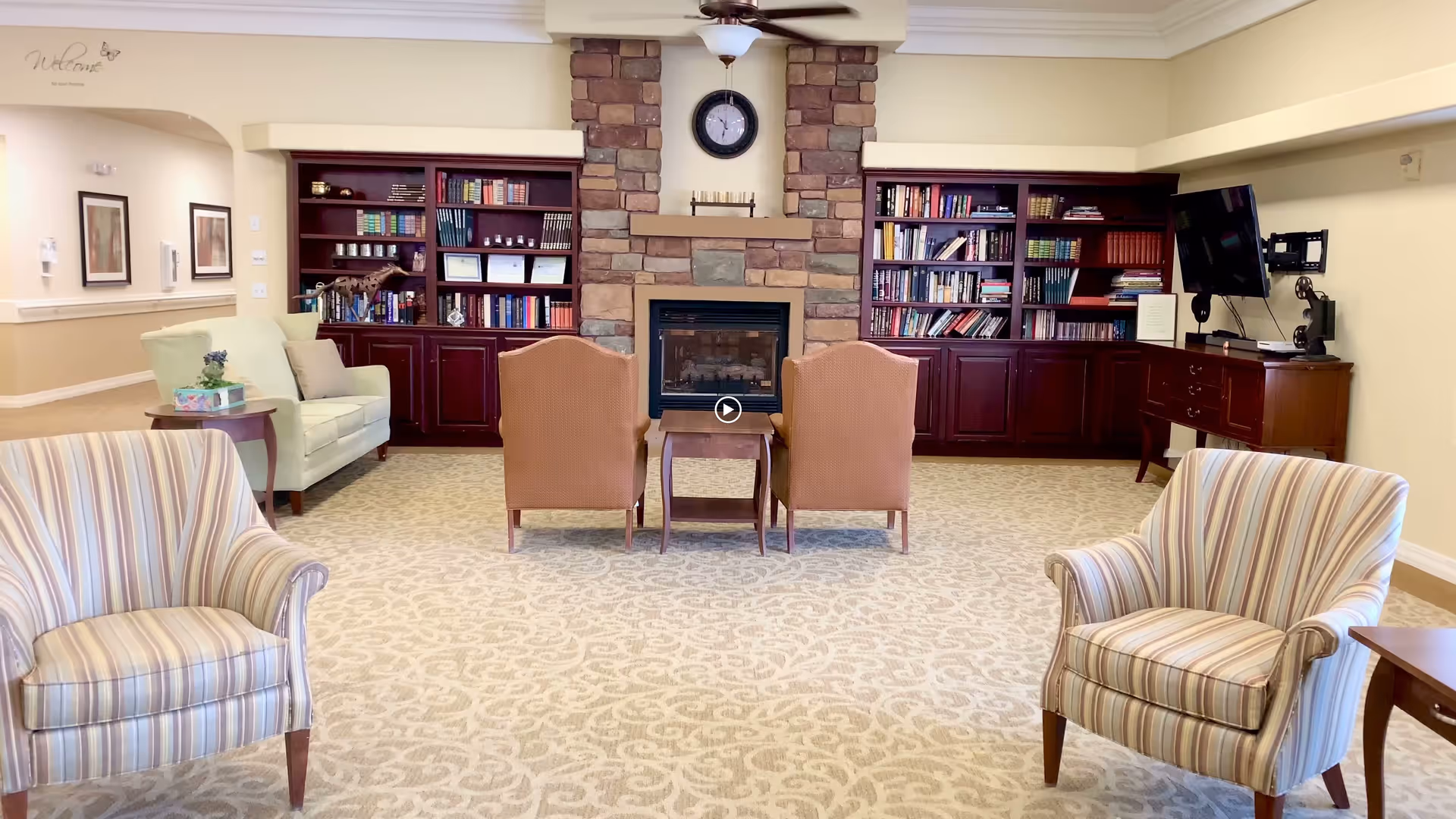 A cozy living room area with two striped armchairs in the foreground, two brown chairs facing a stone fireplace with a clock above it, and built-in dark wood bookshelves filled with books on either side of the fireplace. There is a light green armchair with a side table on the left and a TV mounted on a wooden cabinet on the right. The room has beige patterned carpet and cream-colored walls.