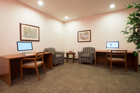 Small interior computer lounge with two desks each holding a desktop computer, two upholstered armchairs around a small table, framed artwork on the walls, and a potted plant.