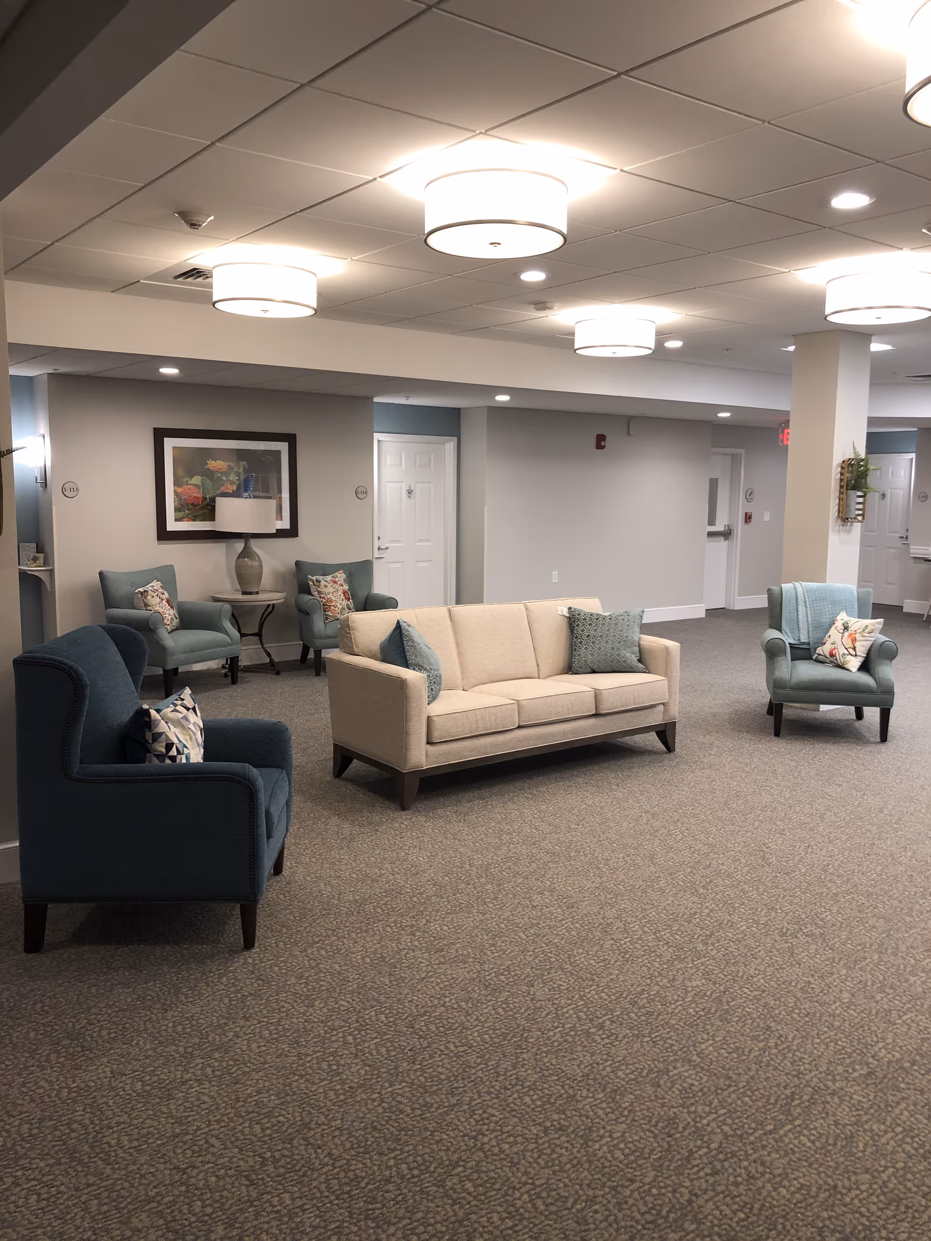 A spacious and well-lit common area with a beige sofa and three armchairs in shades of blue and teal, each with decorative pillows. The room has a carpeted floor, white ceiling with round light fixtures, and light gray walls. There is a framed floral picture on the wall and several doors visible in the background.