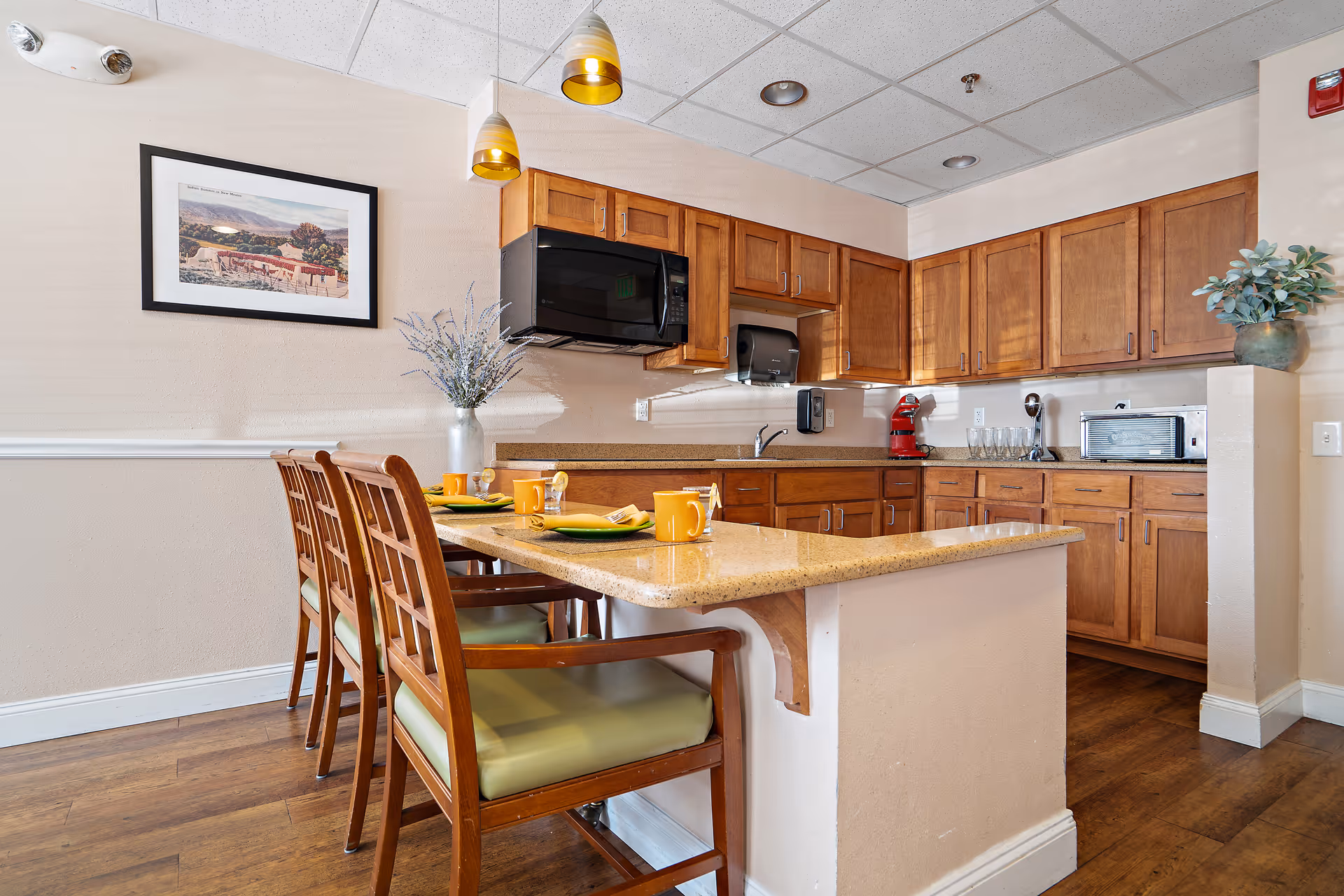 A kitchen area with wooden cabinets, a microwave, a toaster oven, and a red stand mixer. There is a countertop with four wooden chairs with green cushions, set with yellow mugs and green plates. A vase with flowers is placed on the counter. The walls are beige with a framed picture hanging on one side.