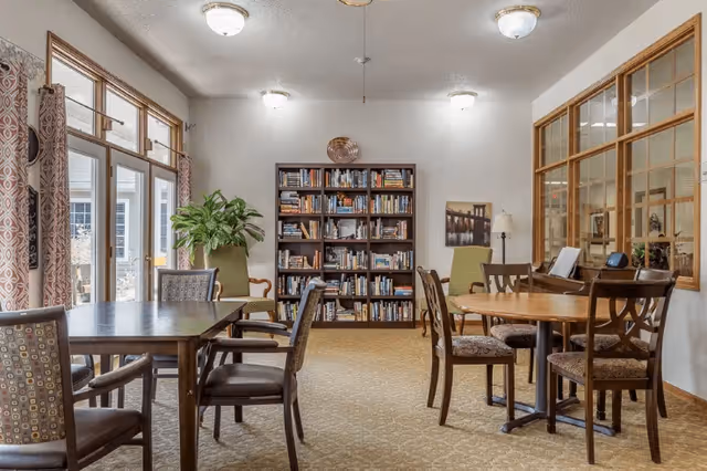 A cozy interior room with multiple wooden tables and chairs arranged for seating. A tall bookshelf filled with books is against the back wall, flanked by two green armchairs. Large windows with patterned curtains allow natural light to enter the room. The walls are light-colored, and there is a framed picture and a floor lamp near the bookshelf.