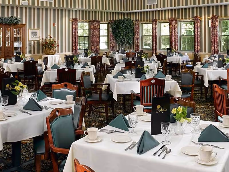 A formal dining room with multiple tables covered in white tablecloths, each set with green folded napkins, cups, plates, glasses, and silverware. The room features wooden chairs with green cushions, striped wallpaper, floral curtains, and large windows letting in natural light. Menus with the Atria logo are placed on several tables.