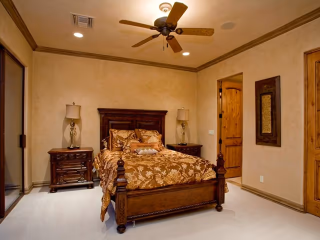 Warmly lit bedroom with a carved wooden bed, patterned bedding, matching nightstands and lamps, a ceiling fan, and neutral walls.