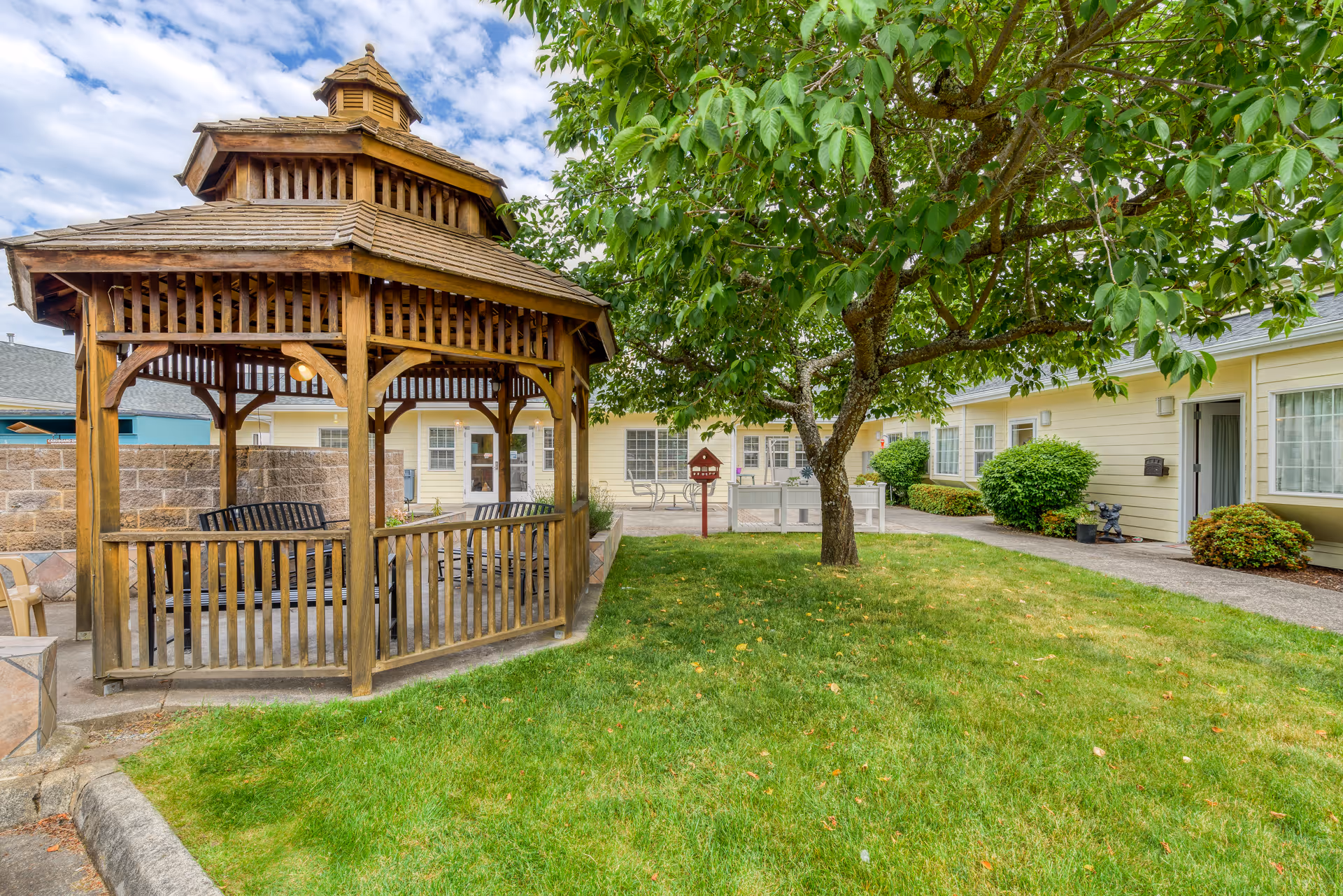 Outdoor garden area at Easthaven Villa Assisted Living & Memory Care featuring a wooden gazebo with benches, a green lawn, a tree with leafy branches, and a yellow building with multiple windows and doors in the background.