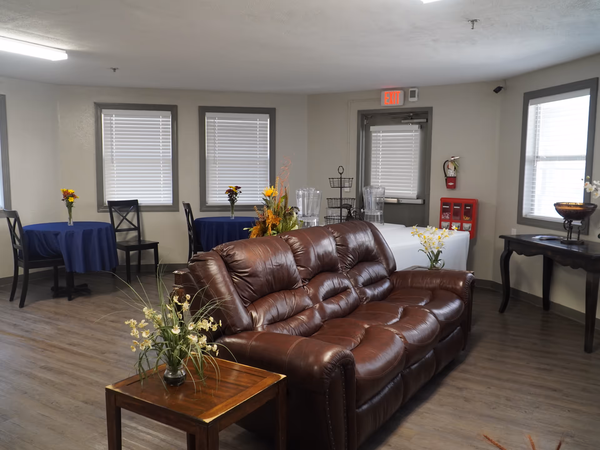 Interior view of a common area in Canterbury House Assisted Living featuring a brown leather sofa, small tables with blue tablecloths and flower arrangements, a wooden side table with a plant, and windows with blinds. There is a door with an exit sign above it and a fire extinguisher mounted on the wall.