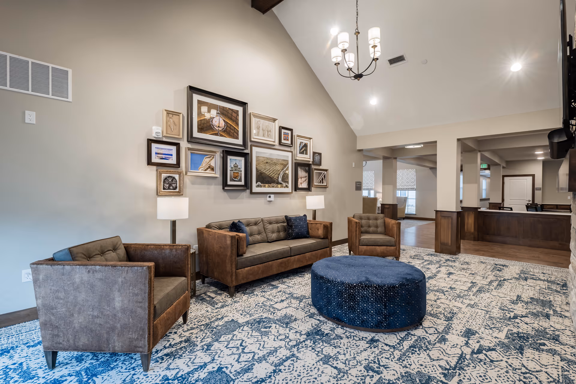 A spacious senior living facility lounge area with a high vaulted ceiling, featuring a brown leather sofa, two matching armchairs, and a large round blue ottoman on a patterned blue and white rug. The wall behind the sofa is decorated with a gallery of framed pictures. Two floor lamps flank the sofa, and a chandelier hangs from the ceiling. The room opens into another area with wooden paneling and additional seating.