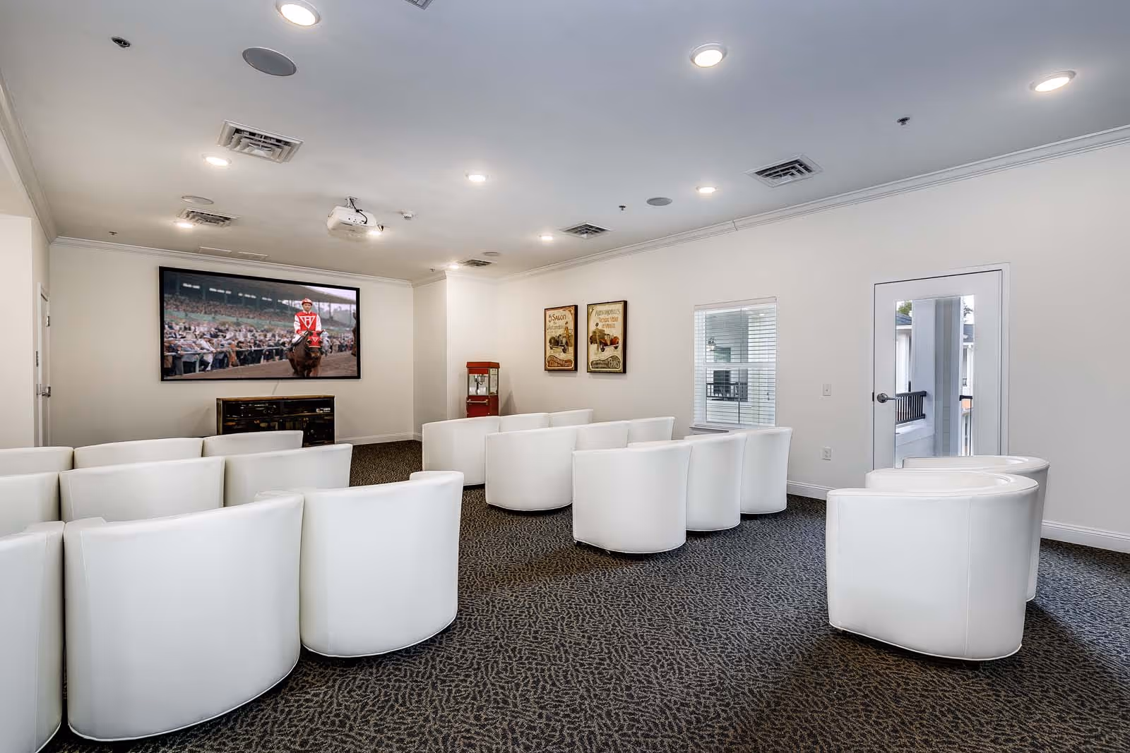 A small theater room with white curved chairs arranged in rows facing a large screen displaying a horse race. The room has white walls, a carpeted floor, recessed ceiling lights, a popcorn machine in the corner, and two vintage-style posters on the wall.