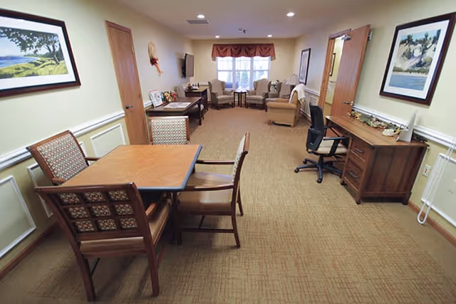 A cozy interior room with beige carpet and cream-colored walls featuring a wooden table with four chairs in the foreground. In the background, there are two armchairs and a loveseat arranged near a window with a valance. A flat-screen TV is mounted on the wall, and a wooden desk with an office chair and computer is positioned on the right side. The room is decorated with framed landscape paintings and small decorative items.