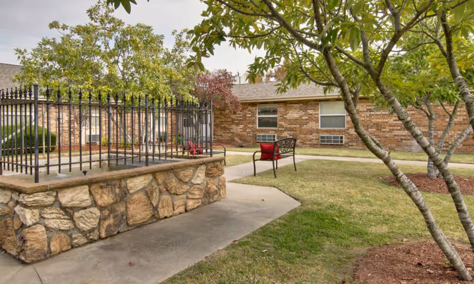 A courtyard with a stone planter topped by a metal fence, a bench with red cushions, trees, and a one-story brick building in the background.