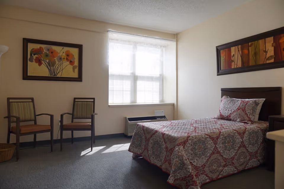 A simple bedroom with a single bed covered in a patterned quilt, two wooden chairs with cushions, a window with blinds letting in natural light, and two framed floral paintings on the walls.