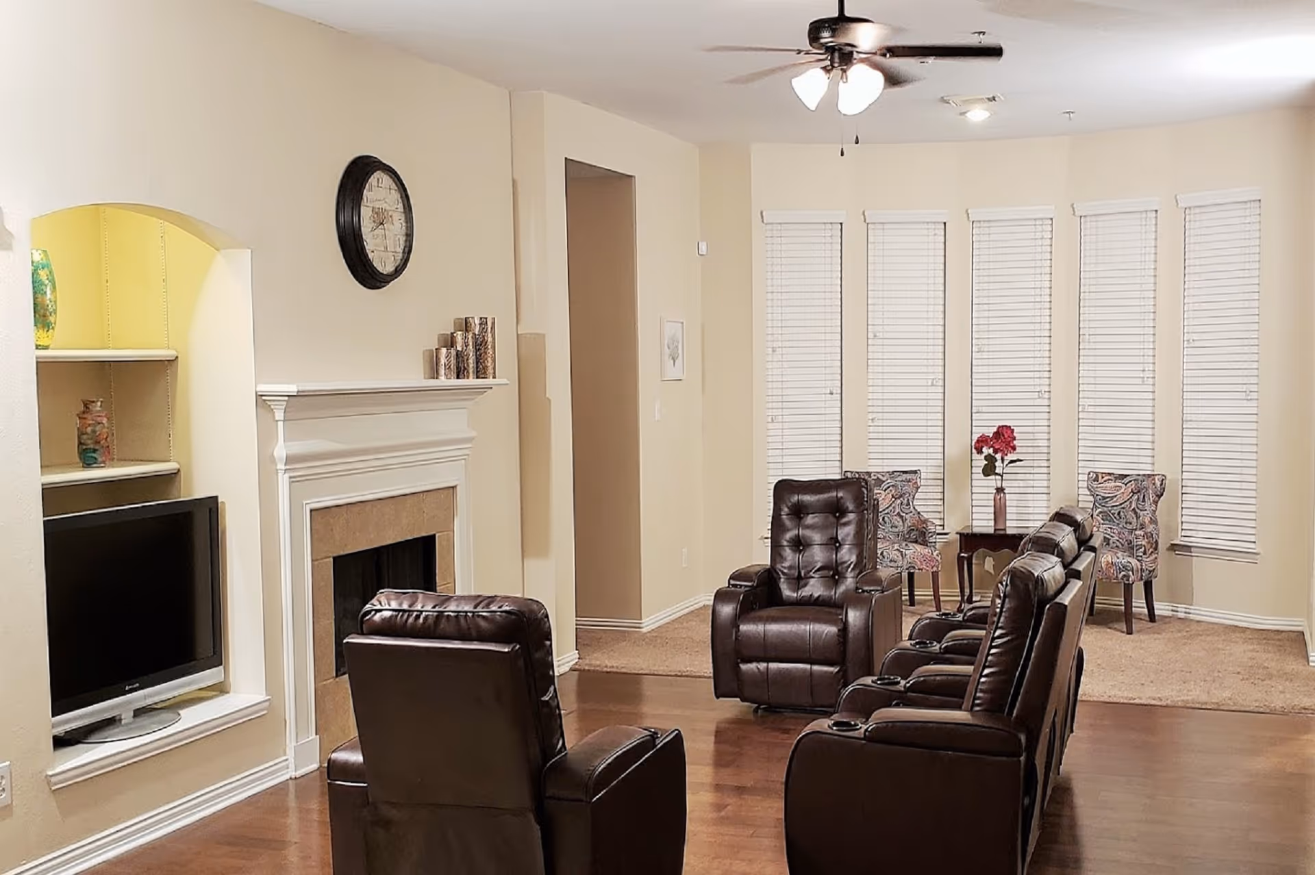 A cozy living room with four brown leather recliners arranged facing a fireplace and a flat-screen TV mounted in a recessed wall niche. The room features light beige walls, a ceiling fan with lights, and a set of five tall windows with white blinds. In the corner, there is a small table with a vase of red flowers and two patterned chairs. The floor is a combination of wood and carpet.