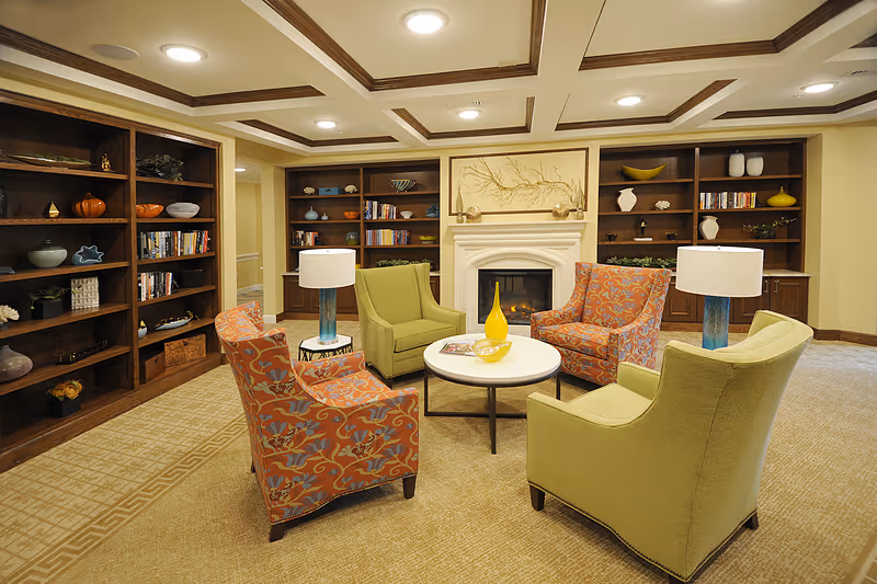A cozy senior living common area with four armchairs arranged around a round white coffee table. Two chairs are green and two have an orange floral pattern. The room features built-in wooden bookshelves filled with books and decorative items, a fireplace with a framed artwork above it, and two table lamps with blue bases on either side of the seating area. The ceiling has recessed lighting and wooden trim, and the floor is carpeted.