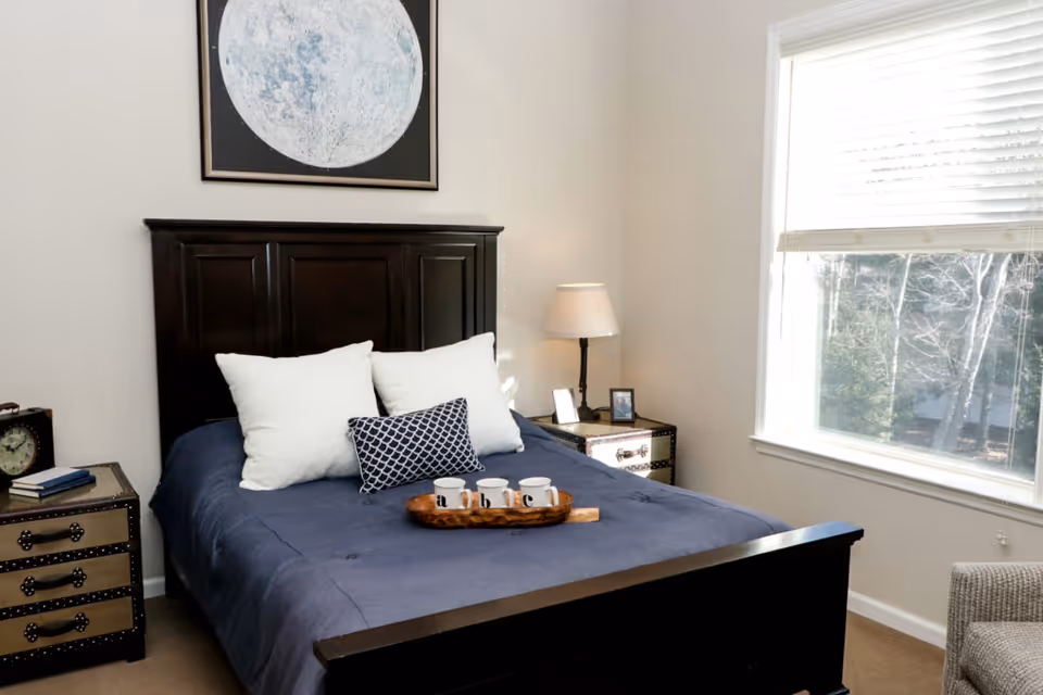 A bedroom with a dark wooden bed frame and a blue bedspread, decorated with three pillows including two white and one patterned. On the bed is a wooden tray holding three white mugs. There are two matching nightstands on either side of the bed, one with a lamp and framed photos, and the other with a clock and books. A large window with white blinds allows natural light to fill the room. Above the bed hangs a framed picture of the moon.