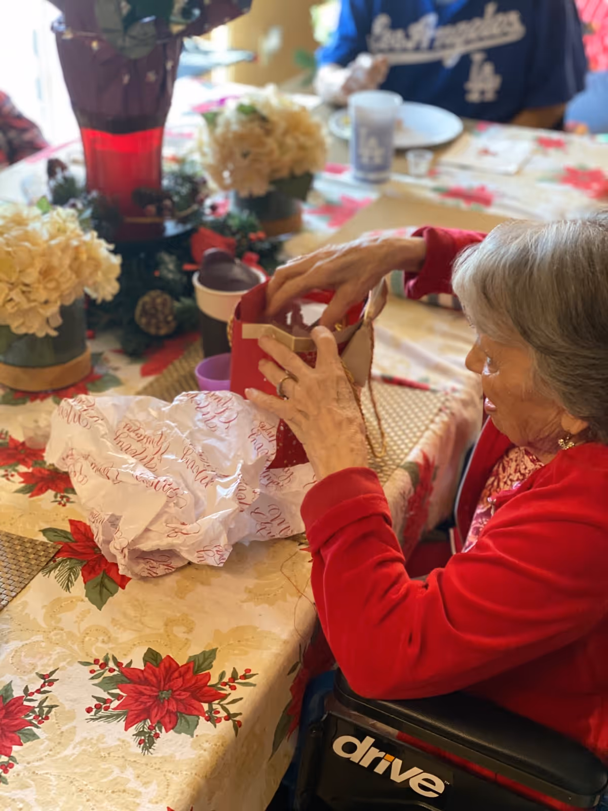 An elderly woman in a red sweater sitting in a wheelchair at a table decorated with a festive poinsettia tablecloth. She is reaching into a red gift bag with tissue paper on the table. There are floral centerpieces and other people partially visible in the background.
