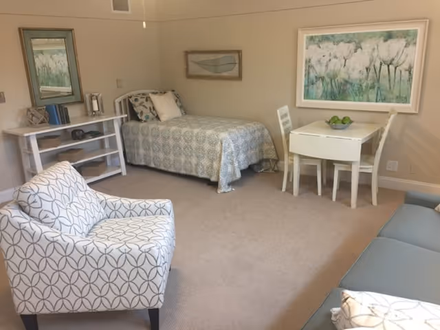A cozy room with a single bed covered in a patterned quilt, a small white table with two chairs, a cushioned armchair with a geometric pattern, a blue sofa, and framed artwork on the walls. There is also a white shelving unit with books and decorative items.