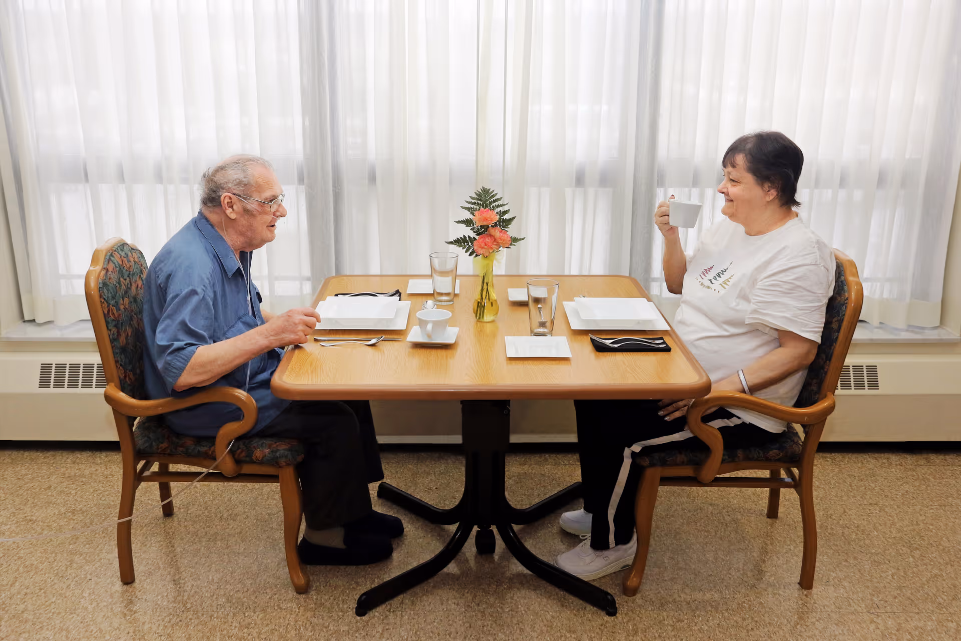 An elderly man and woman sitting across from each other at a wooden dining table with place settings, glasses, and a small vase with flowers in the center. The man is wearing a blue shirt and glasses, and the woman is wearing a white t-shirt and holding a white cup. They are in a room with large windows covered by sheer white curtains.