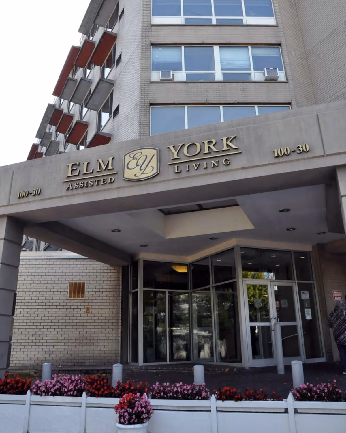 Entrance of Elm York Assisted Living facility showing the building's name above the doorway, glass doors, and a flower bed with red and pink flowers in front.