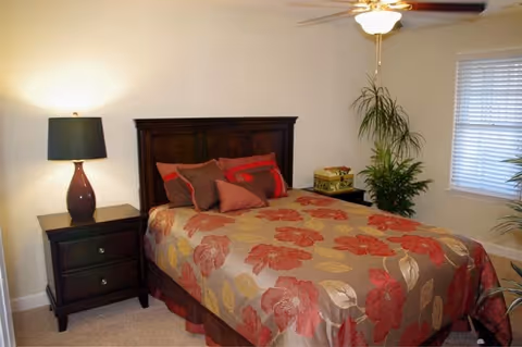 A bedroom with a bed featuring a floral bedspread in shades of red, beige, and brown. The bed has multiple pillows in coordinating colors. To the left of the bed is a dark wooden nightstand with two drawers and a lamp with a dark base and light shade. There is a ceiling fan with a light fixture above the bed. A window with white blinds is on the right wall, and there are green plants near the window and nightstand. The walls are painted light beige.
