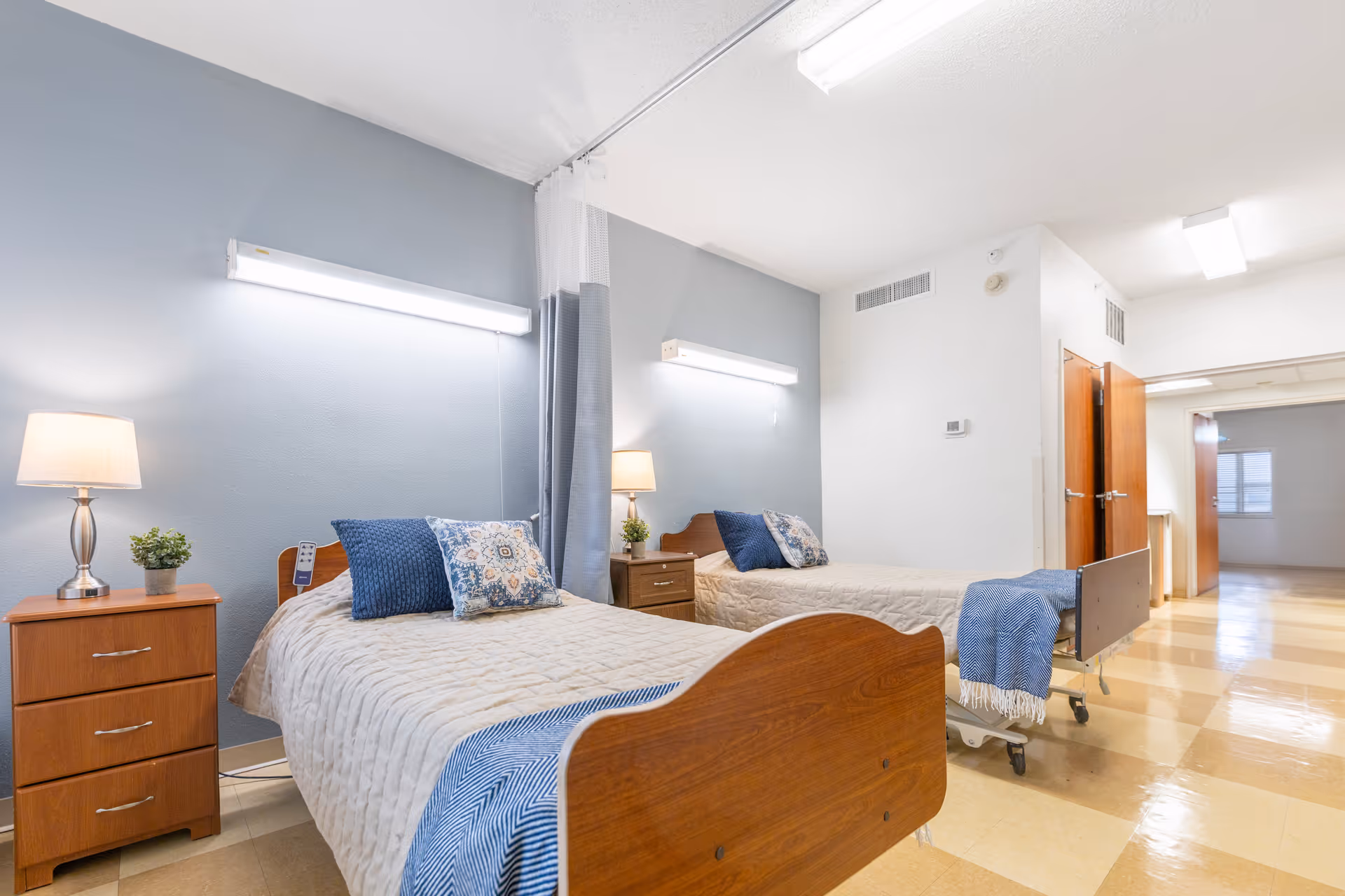 A bright nursing facility room with two single beds, each with beige bedspreads, blue and patterned pillows, and blue throws at the foot. Each bed has a wooden nightstand with a lamp and a small plant. The walls are painted light blue and white, and there is a privacy curtain between the beds. The floor has a checkered pattern in beige tones, and the room is well-lit with ceiling lights.
