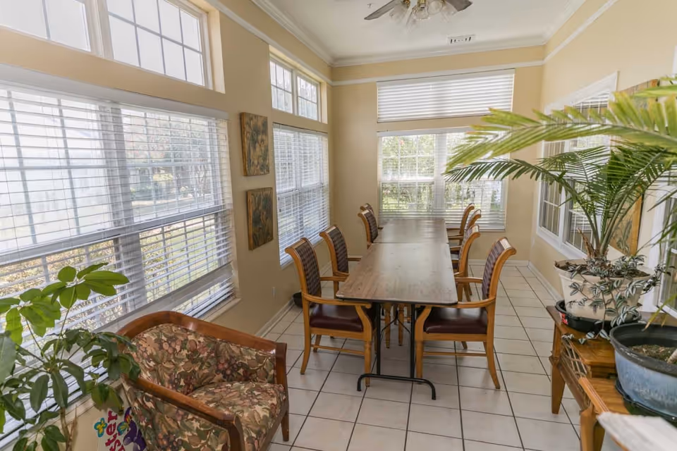 A bright dining room with a long wooden table surrounded by eight wooden chairs with cushioned seats. The room has large windows with white blinds letting in natural light. There are two floral paintings on the wall, a floral upholstered armchair, and several potted plants adding greenery to the space. The floor is tiled in white.