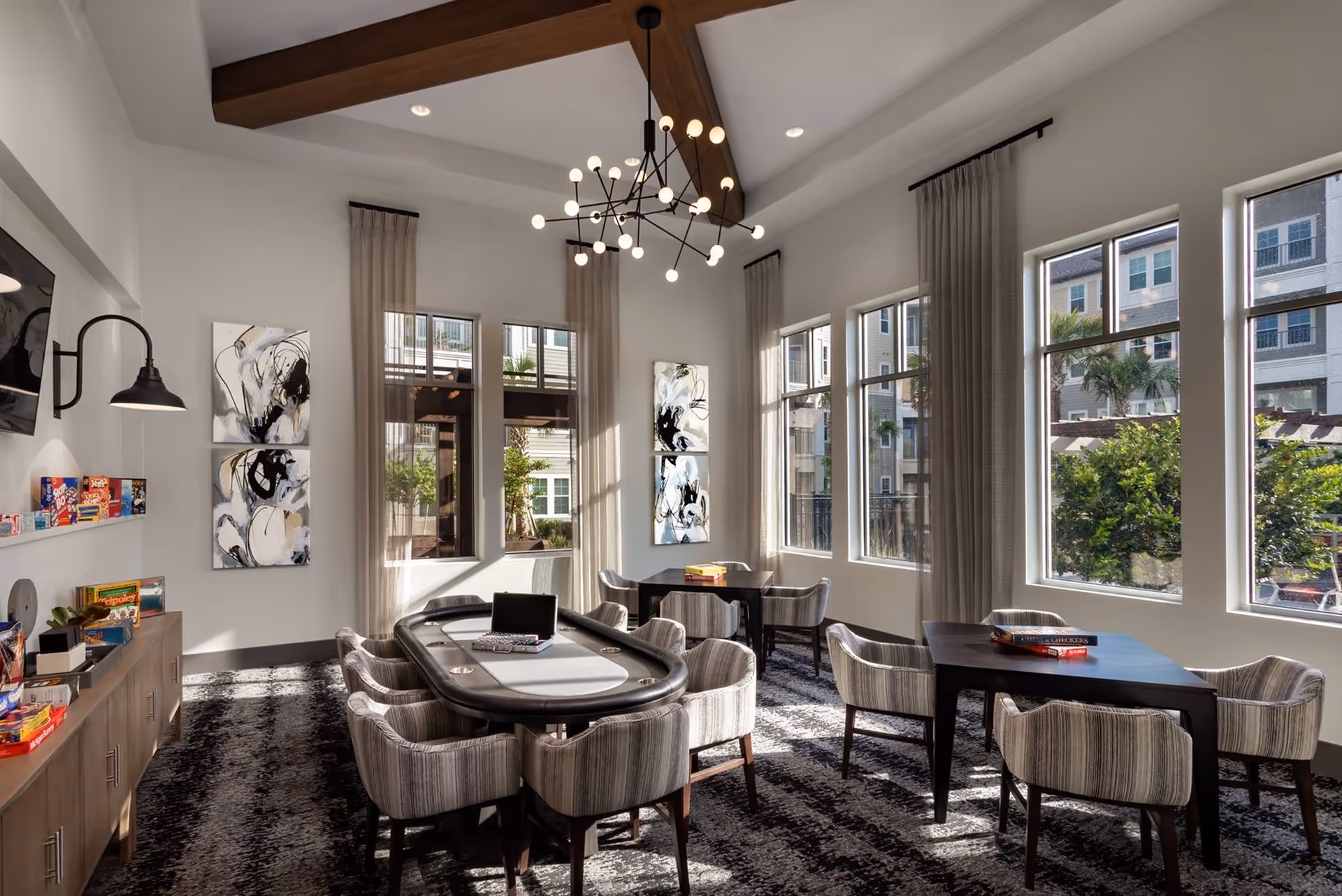 A bright and spacious game room with large windows letting in natural light. The room features a poker table surrounded by cushioned chairs, additional tables with chairs, modern abstract wall art, and a contemporary chandelier hanging from a ceiling with exposed wooden beams. Outside the windows, parts of a residential building and greenery are visible.