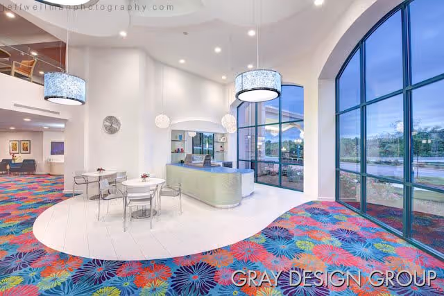 Bright and modern interior space with large arched windows, colorful floral carpet, a reception desk, round tables with chairs, and contemporary pendant lights hanging from the ceiling.