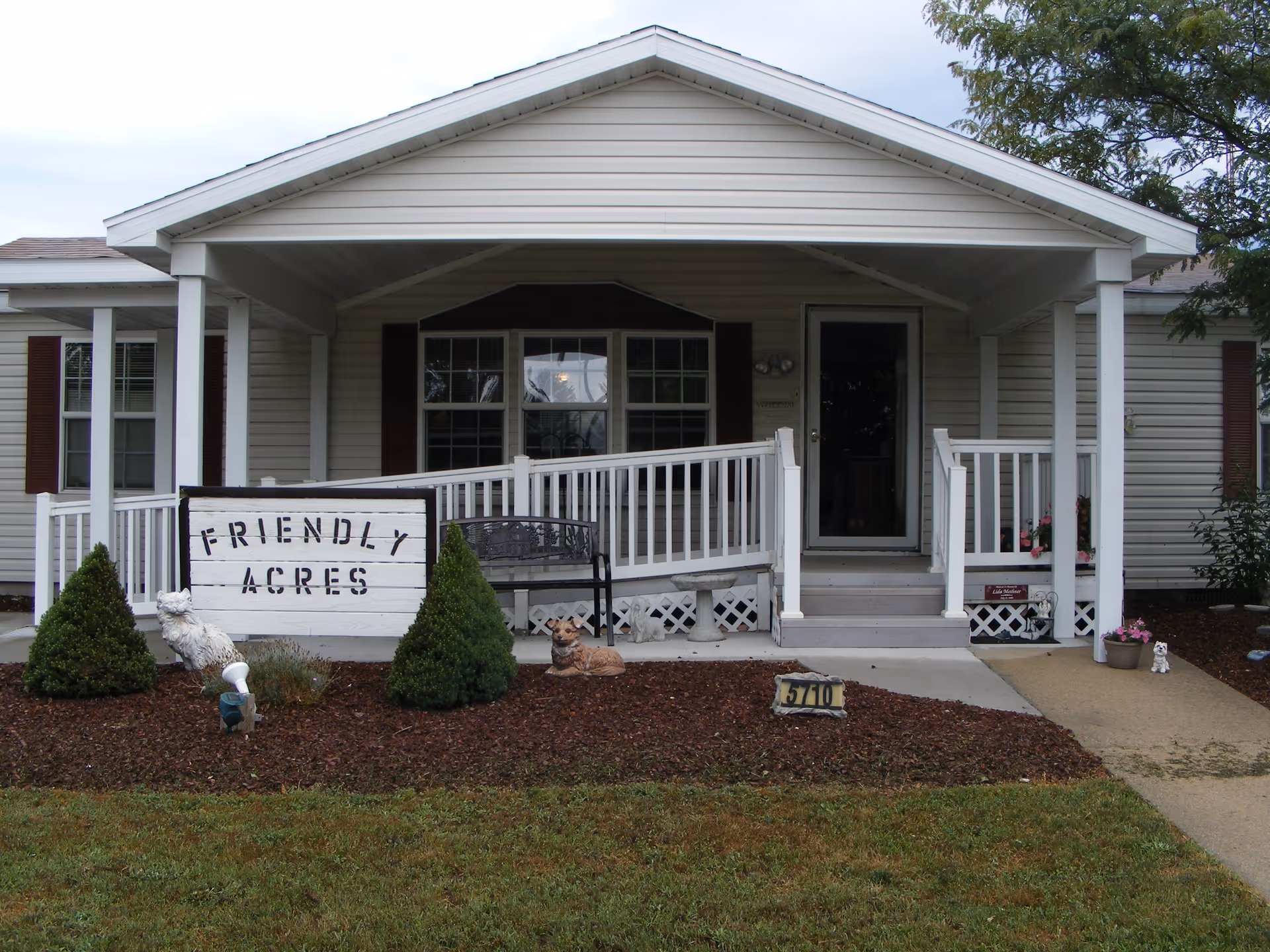 Front exterior view of a single-story building with a covered porch, white railing, and a ramp. There is a sign in the garden bed that reads 'Friendly Acres'. The garden bed has small bushes and decorative animal statues. The building has beige siding and maroon shutters around the windows.