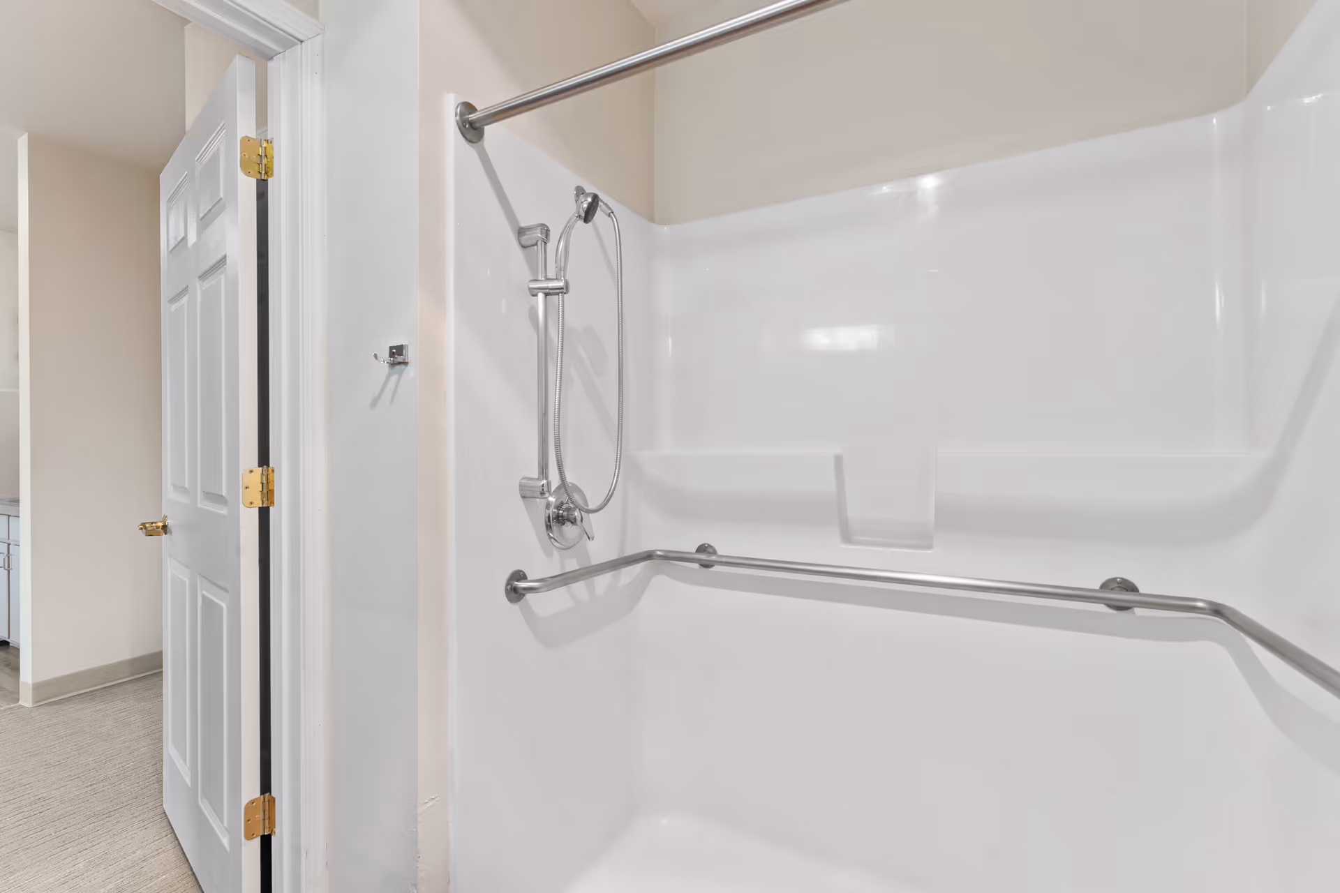 A clean, white shower stall with a handheld showerhead and a horizontal grab bar installed on the wall. The shower is adjacent to a partially open white door leading to a hallway with light-colored walls and carpeted flooring.