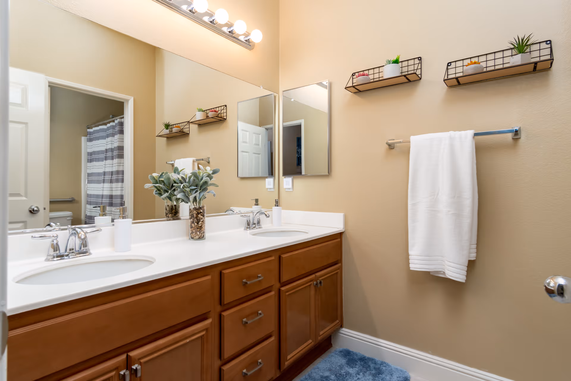 A clean and well-lit bathroom with a double sink vanity featuring wooden cabinets and a white countertop. Above the sinks are two rectangular mirrors and a light fixture with five bulbs. On the countertop, there is a small plant in a glass vase filled with stones and a white soap dispenser. On the beige wall to the right, there are two small black wire shelves holding small potted plants and a white towel hanging on a towel rack. In the background, a shower with a striped curtain is visible through an open door.
