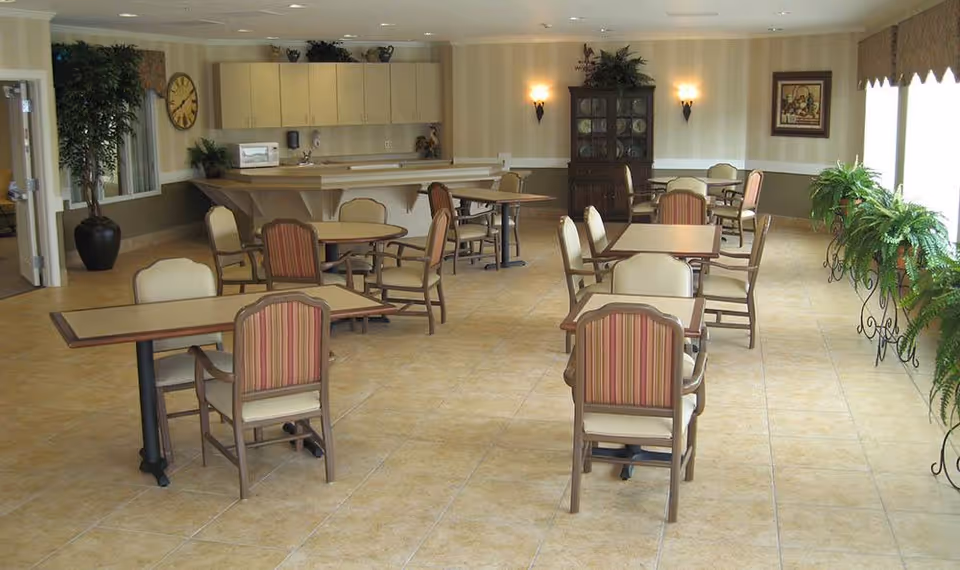 A spacious dining room with multiple tables and chairs arranged neatly. The room features tiled flooring, a large window with potted plants along the windowsill, a wall clock, and a cabinet against the far wall. The lighting is warm with wall sconces and ceiling lights, and there is a counter area with cabinets in the background.