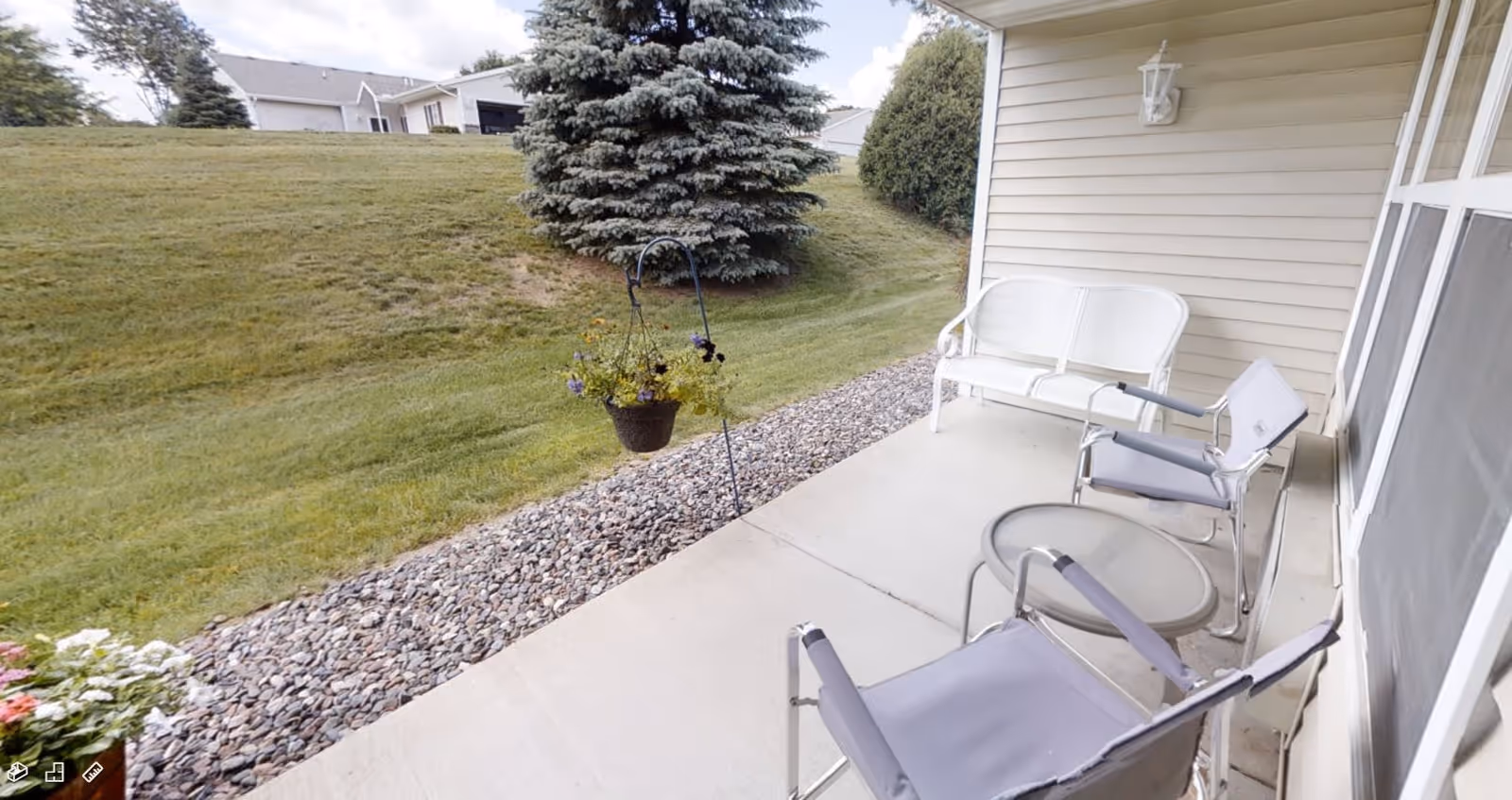 A covered outdoor patio area with two gray chairs, a small round table, and a white bench. The patio overlooks a grassy yard with a large evergreen tree and some houses in the background. There is a hanging flower pot with purple and yellow flowers near the edge of the patio.