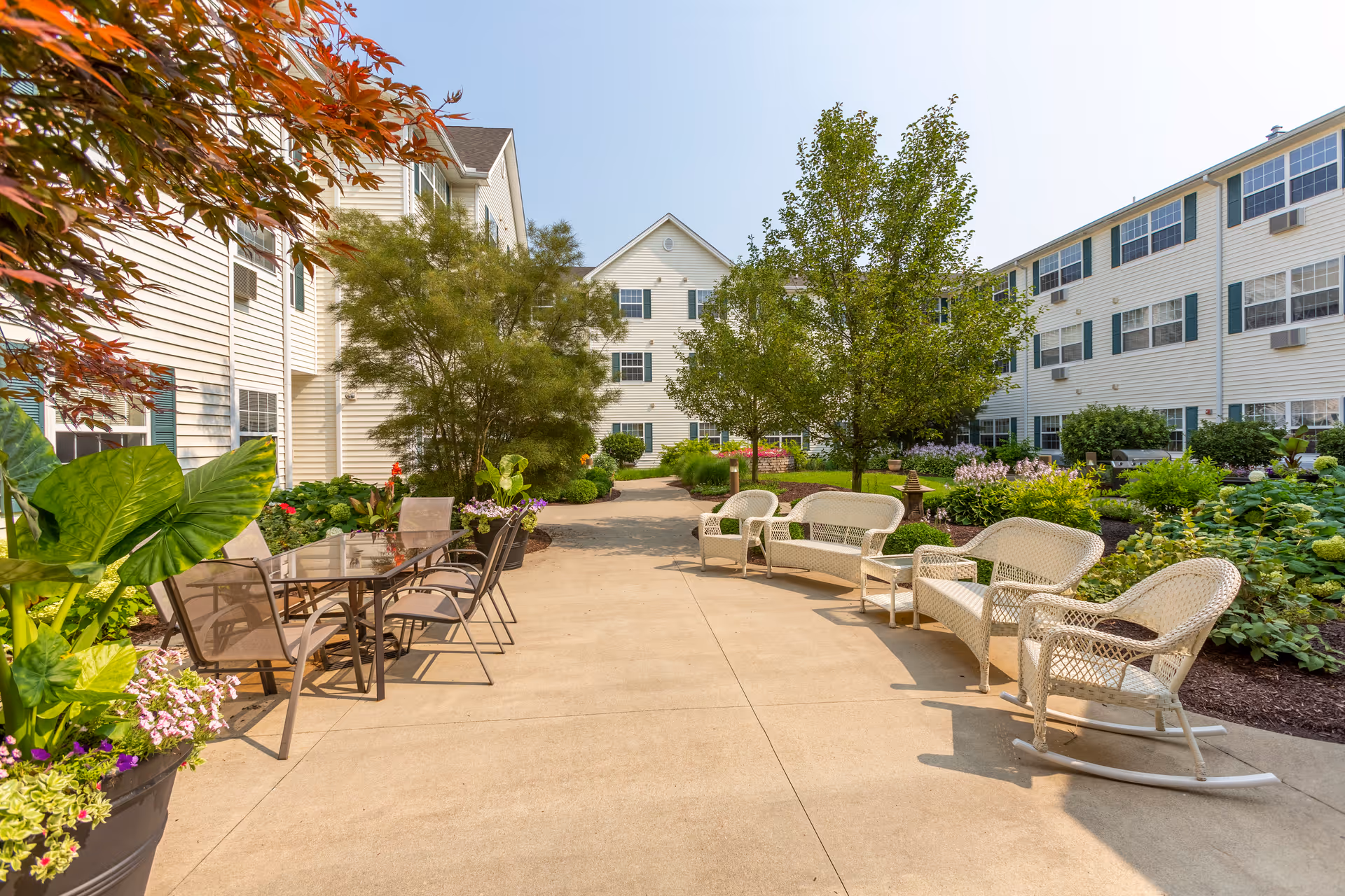 Sunlit courtyard of a senior living facility with wicker chairs, tables, and landscaped gardens between white multi-story buildings.