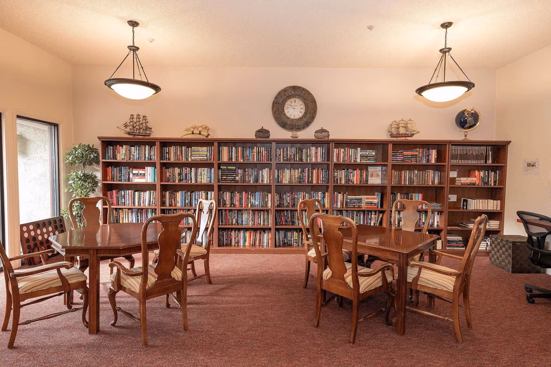 A cozy library room with two wooden tables surrounded by wooden chairs with striped cushions. Behind the tables is a large bookshelf filled with books, decorative ship models, a globe, and a wall clock. The room has warm lighting from two ceiling lamps and a window on the left side letting in natural light.